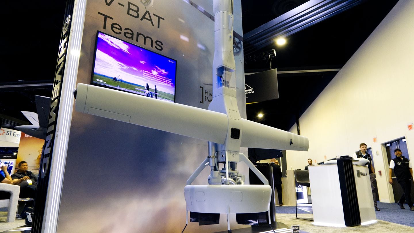 Shield AI's V-Bat drone stands upright at the company's 2024 Sea-Air-Space booth. A screen nearby advertises its swarming capability. (Colin Demarest/C4ISRNET)