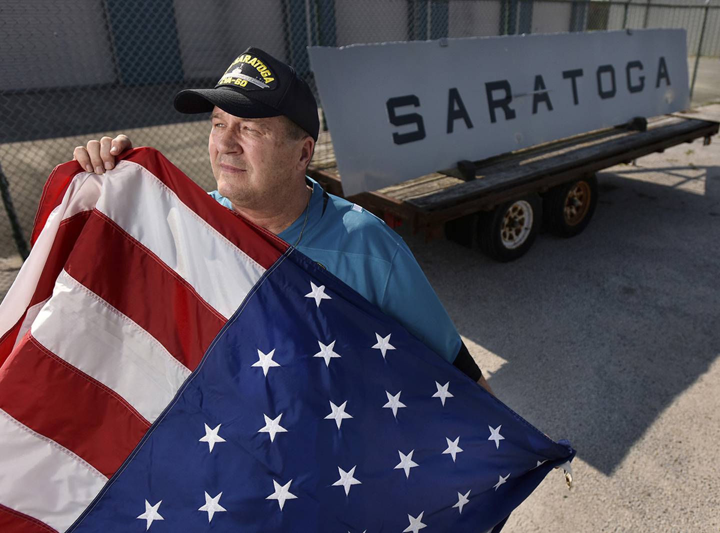 Matt Miller poses with the nameplate of the USS Saratoga on May 29, 2019, while holding the flag he waved when the ship returned from the Persian Gulf in 1991.