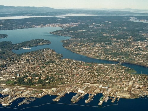 Puget Sound Naval Shipyard and the downtown Bremerton waterfront are viewed from over Port Orchard looking west. The waterways Sinclair Inlet, Port Washington Narrows and Dyes Inlet are visible, with the Olympic Mountains in the background. (Defense Logistics Agency Land and Maritime)